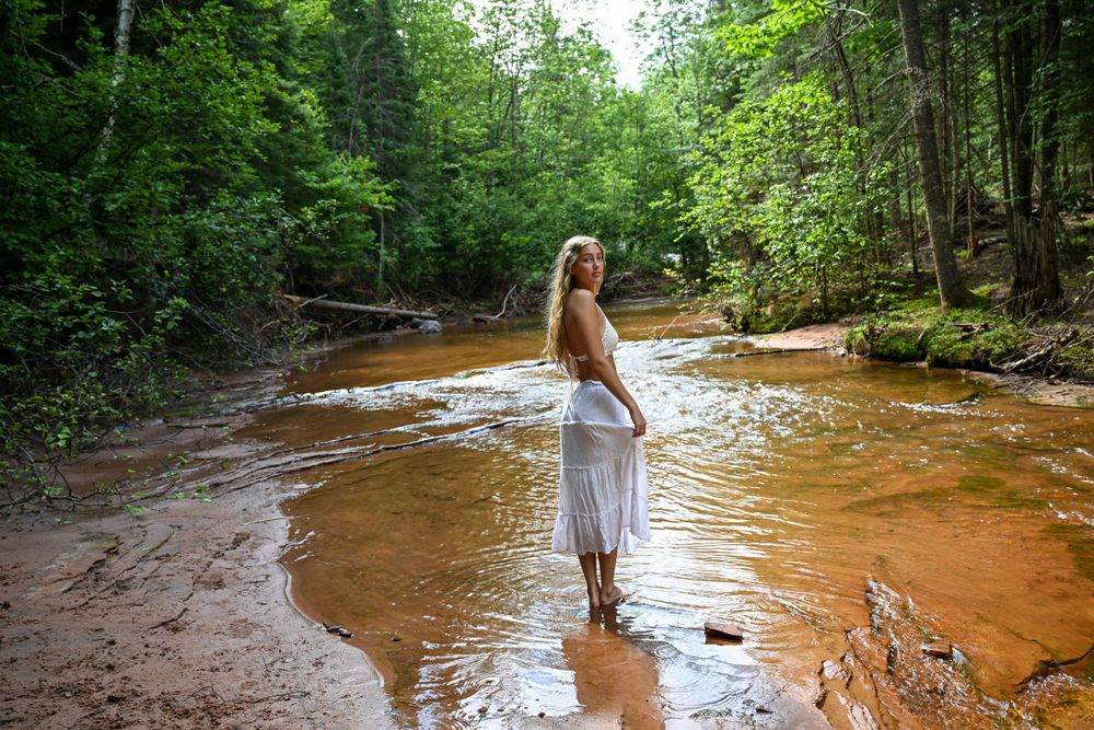 Young woman in white skirt wading in a shallow forest stream with red sandy riverbed.