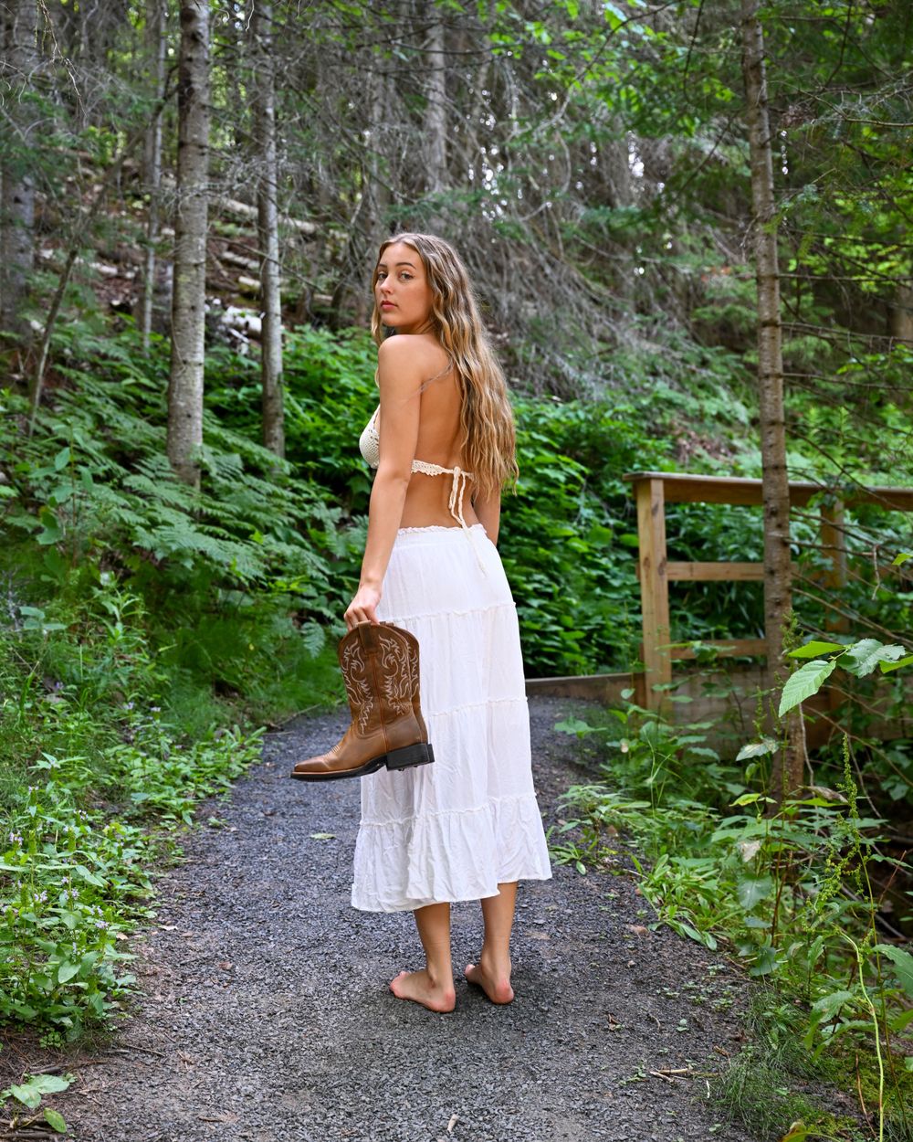 Barefoot woman in white sundress holding cowboy boots on a gravel forest path near wooden gate.