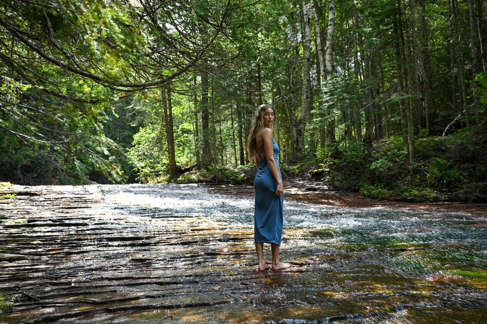 Blonde woman in blue dress standing barefoot in a flowing forest river surrounded by green trees.