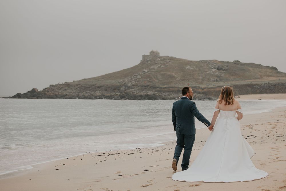 A couple holding hands while walking along a misty beach with a rocky headland in the background at a Cornwall wedding photo shoot