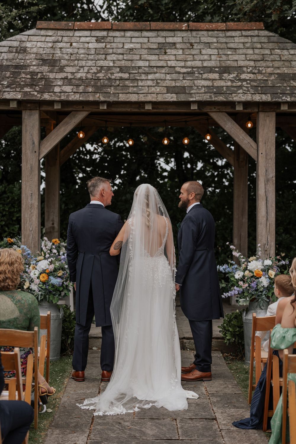 Bride and groom stand at outdoor wooden gazebo altar with floral arrangements during wedding ceremony.