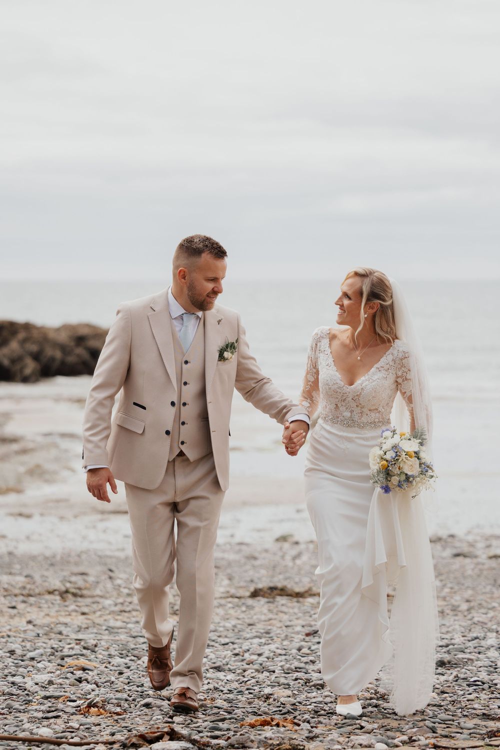 Bride and groom walk hand-in-hand on a rocky beach, she holds a bouquet, he wears a beige suit.