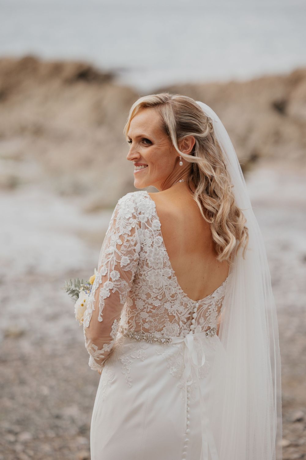 Smiling bride in lace backless wedding dress with veil holding bouquet on beach.