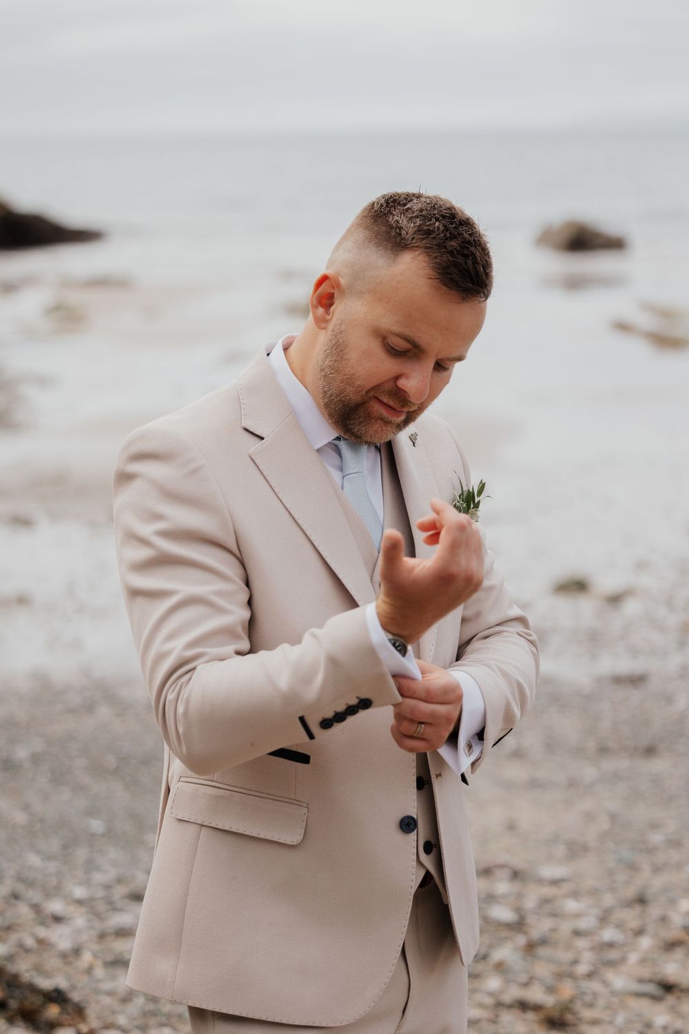 Groom in beige suit adjusting cufflinks on rocky beach with boutonniere.