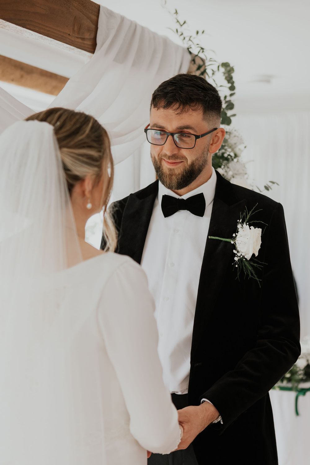 Groom in black tuxedo with bow tie smiles at bride during indoor wedding ceremony.
