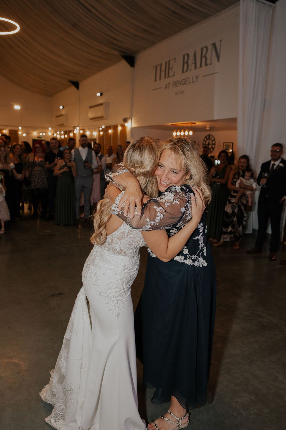 Bride in lace gown hugging guest in navy floral dress at The Barn at Pengelly wedding reception.
