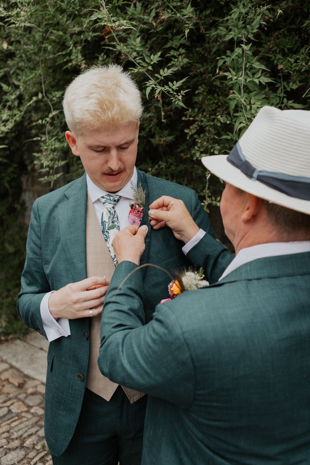 Groom in green suit getting boutonniere pinned by man in hat, outdoor wedding preparation scene.
