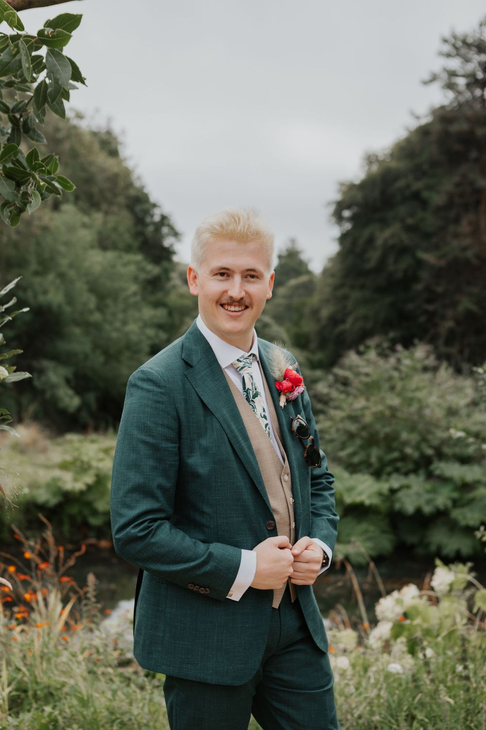 Smiling groom in green suit with red boutonniere and floral tie poses outdoors in lush garden setting.