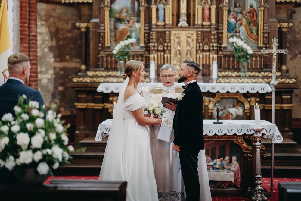 Wedding ceremony taking place in traditional church with red carpet and white floral decorations.