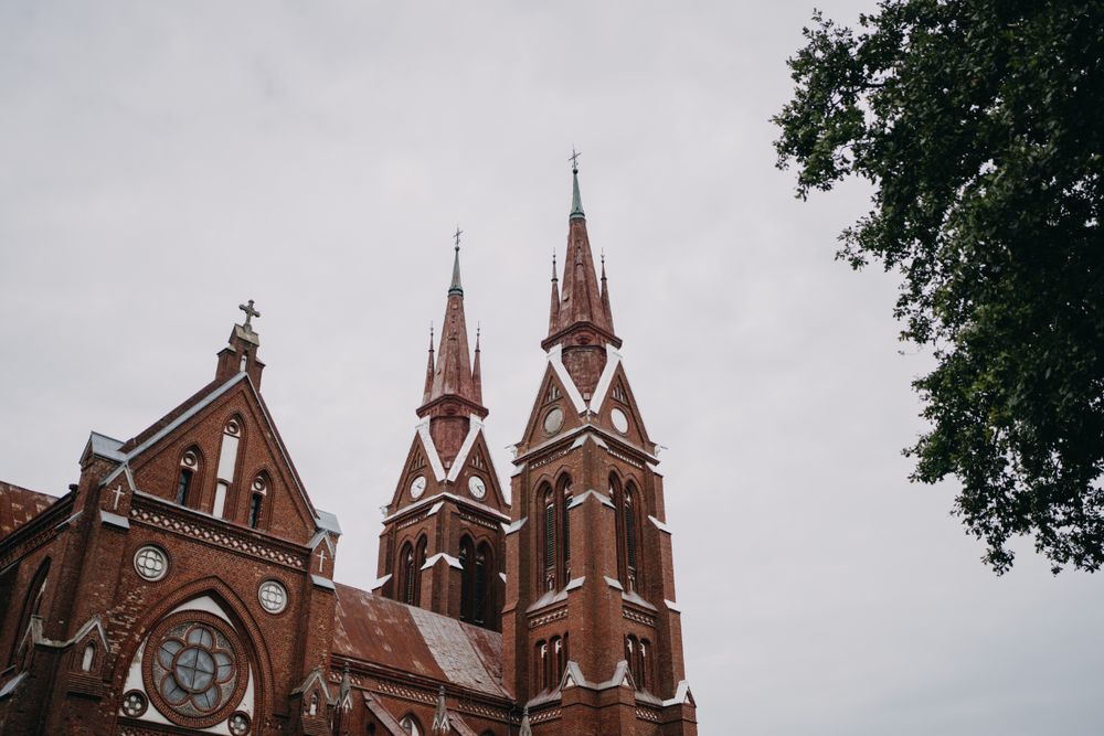 Gothic church exterior with red brick and tall spires against cloudy sky.