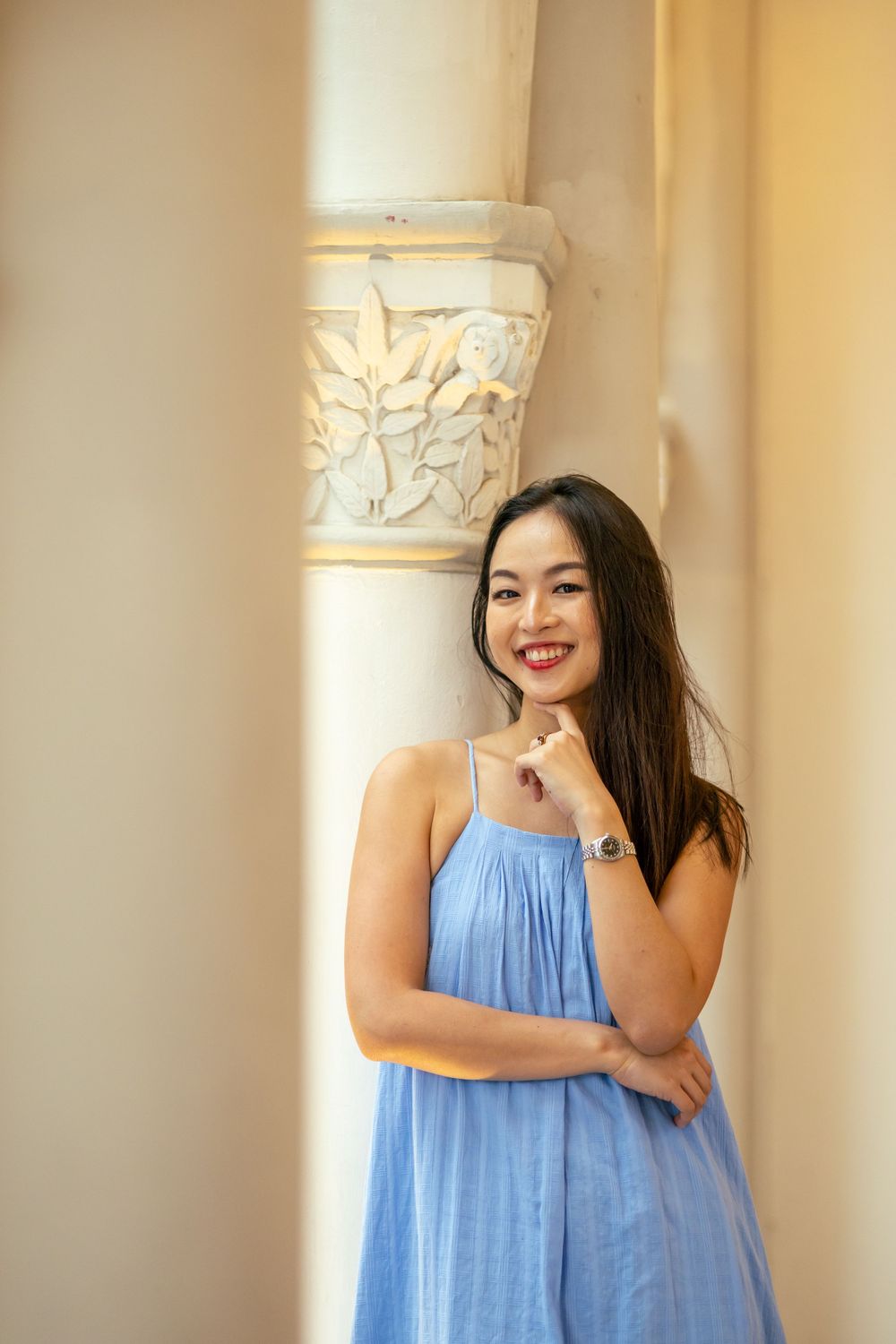 A person in light blue sundress poses against ornate column with carved floral details.