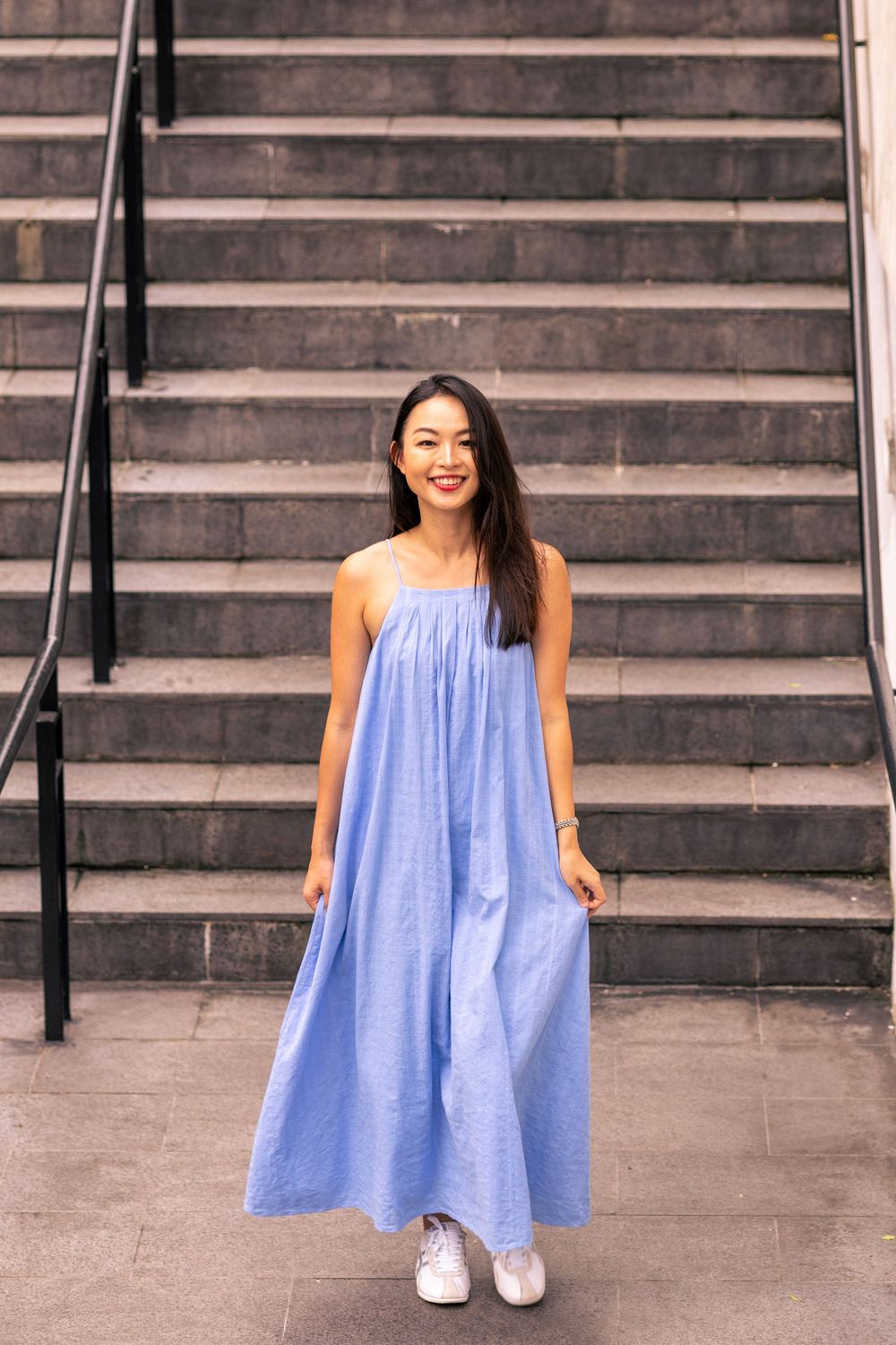 Person in light blue maxi dress stands smiling in front of concrete stairs.