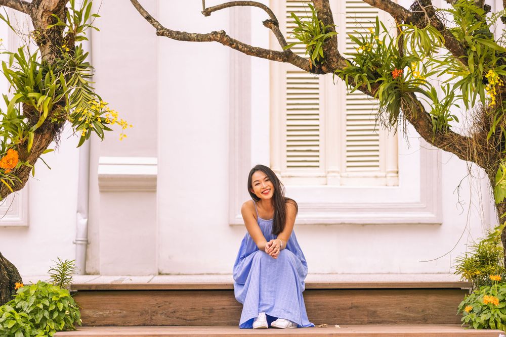 A person in blue dress sits under citrus tree against white colonial building.