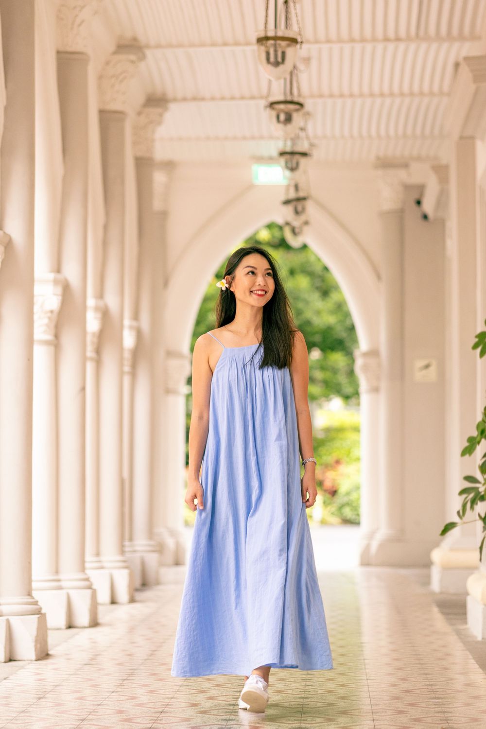 A person in flowing blue maxi dress walks through white colonnade corridor.