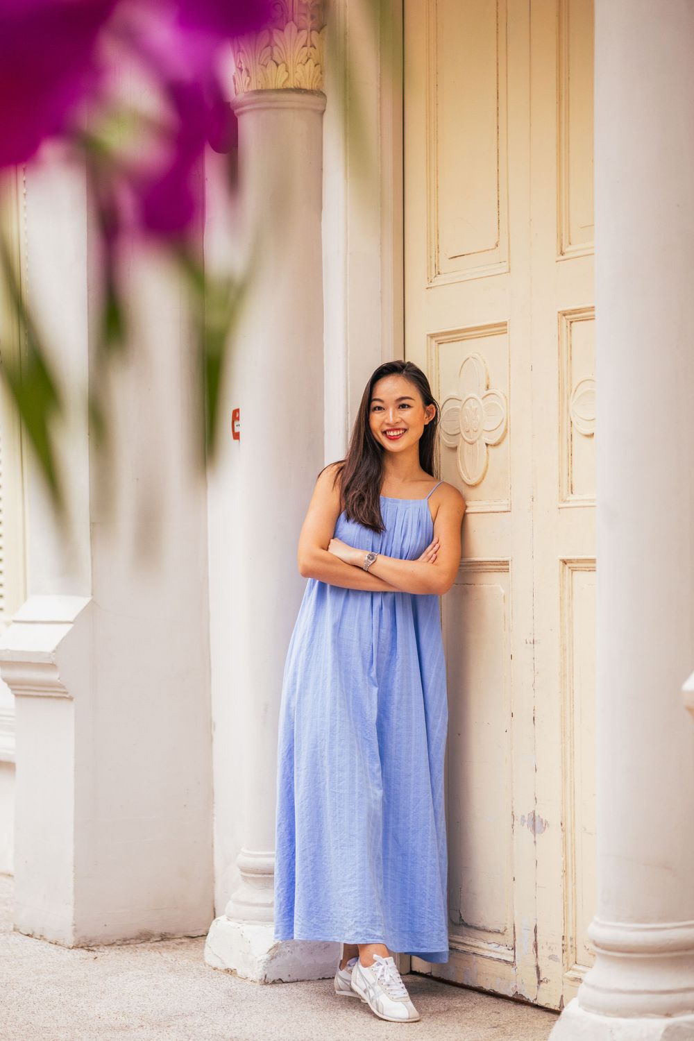 A person in blue maxi dress poses by white colonial pillars with pink flowers.