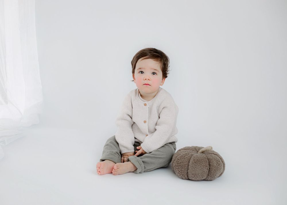 Child sitting with plush toy wearing cream sweater and gray pants on white background.
