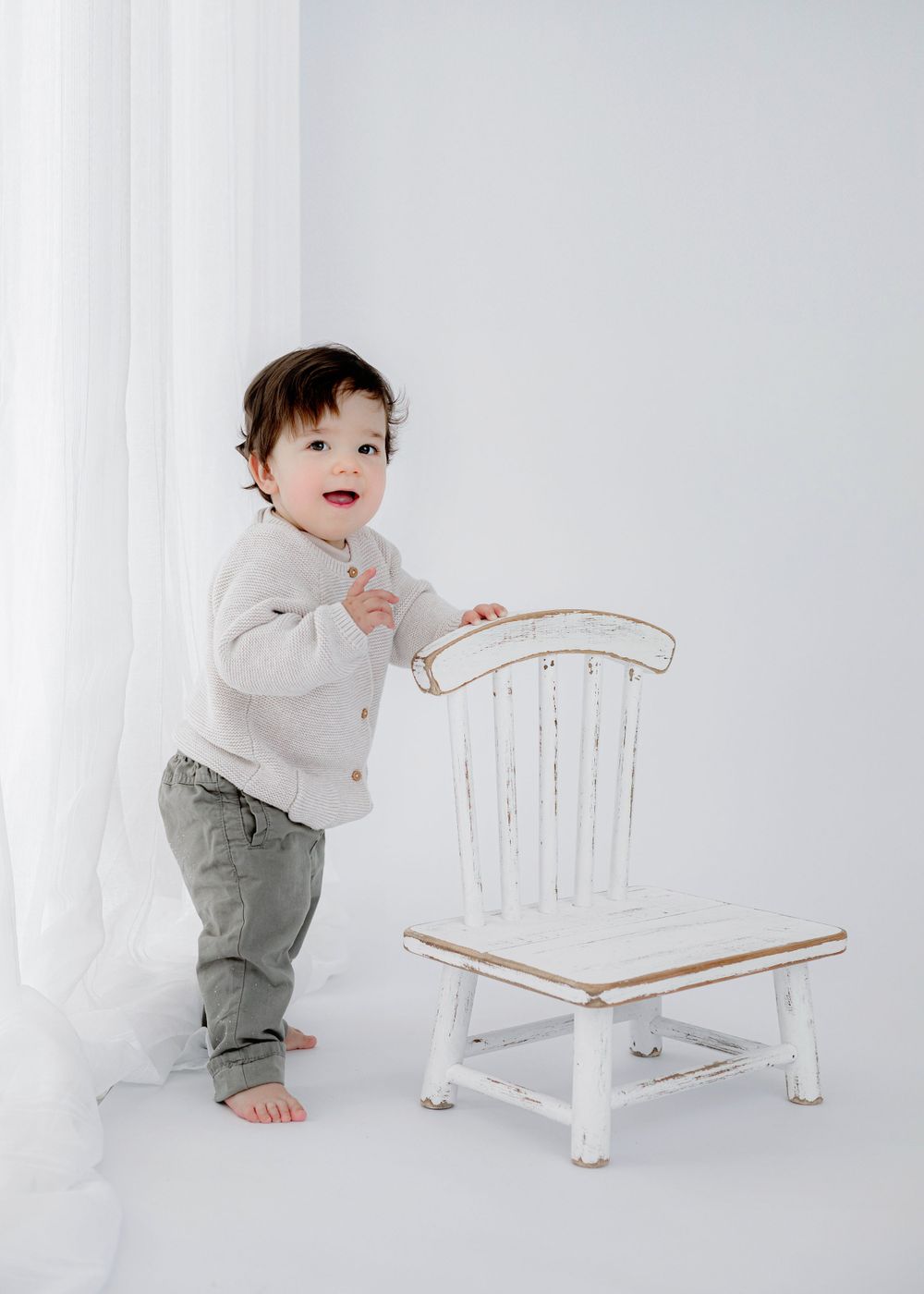 Toddler in gray pants and white cardigan stands barefoot next to vintage white wooden chair against light background.