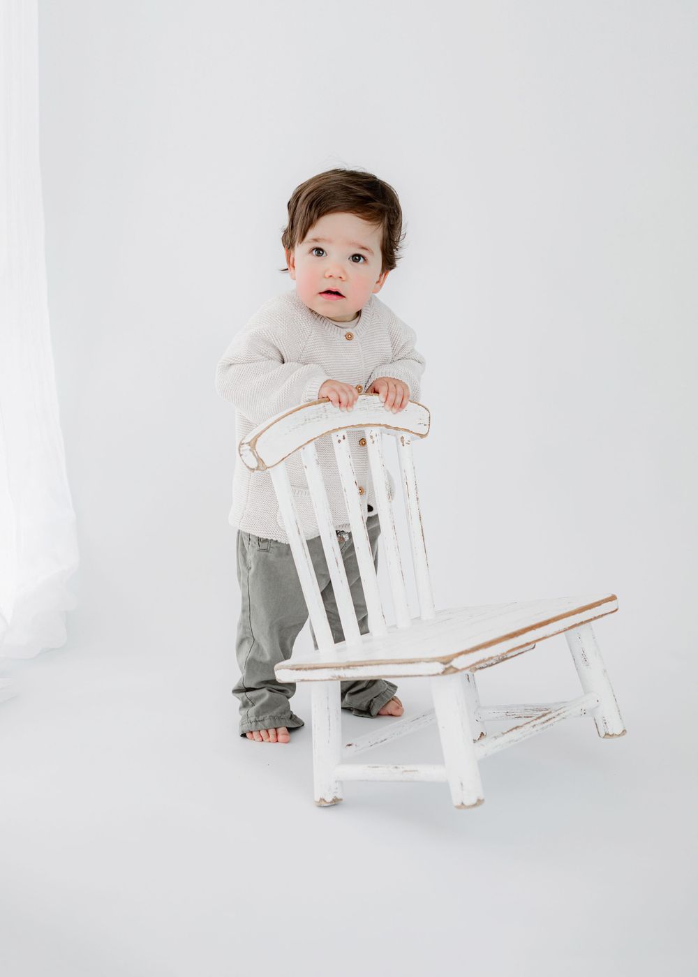 Small child standing next to vintage white chair in minimalist studio setting.