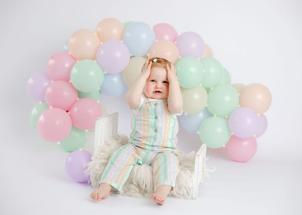Happy baby in ruffled outfit poses with pastel balloon arch and decorative elements.