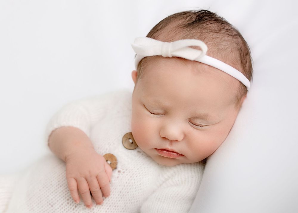 Sleeping newborn baby wearing white bow headband and knit romper on white background.