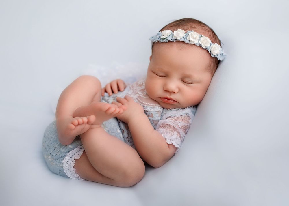 Sleeping baby in delicate lace outfit and floral headband posed on gray background.