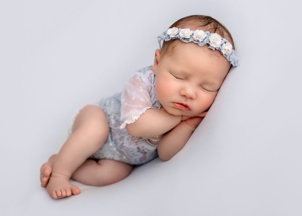 Peaceful sleeping newborn wearing floral headband and lace romper on gray backdrop.