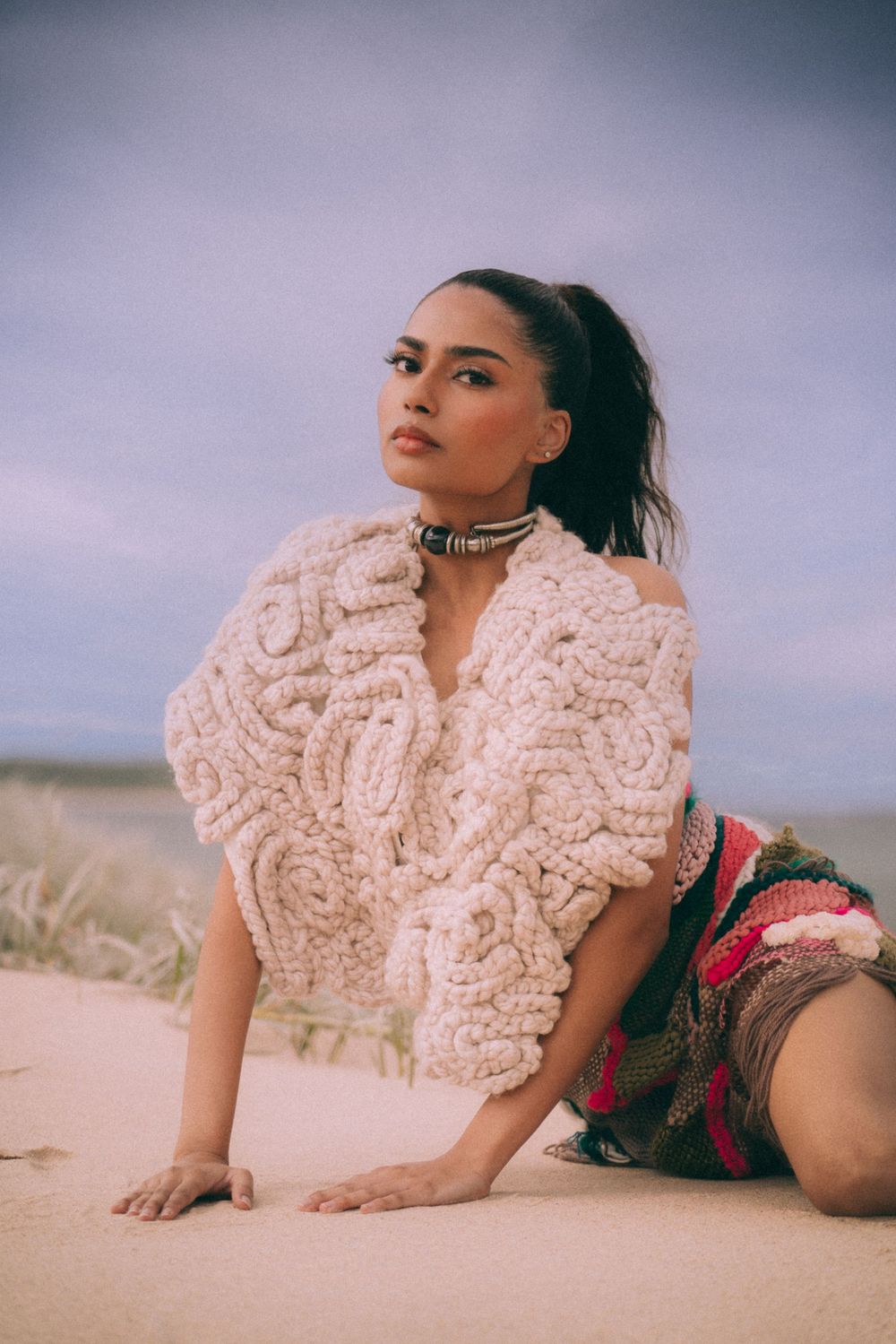 Model in cream textured sweater poses on beach sand during sunset photoshoot.
