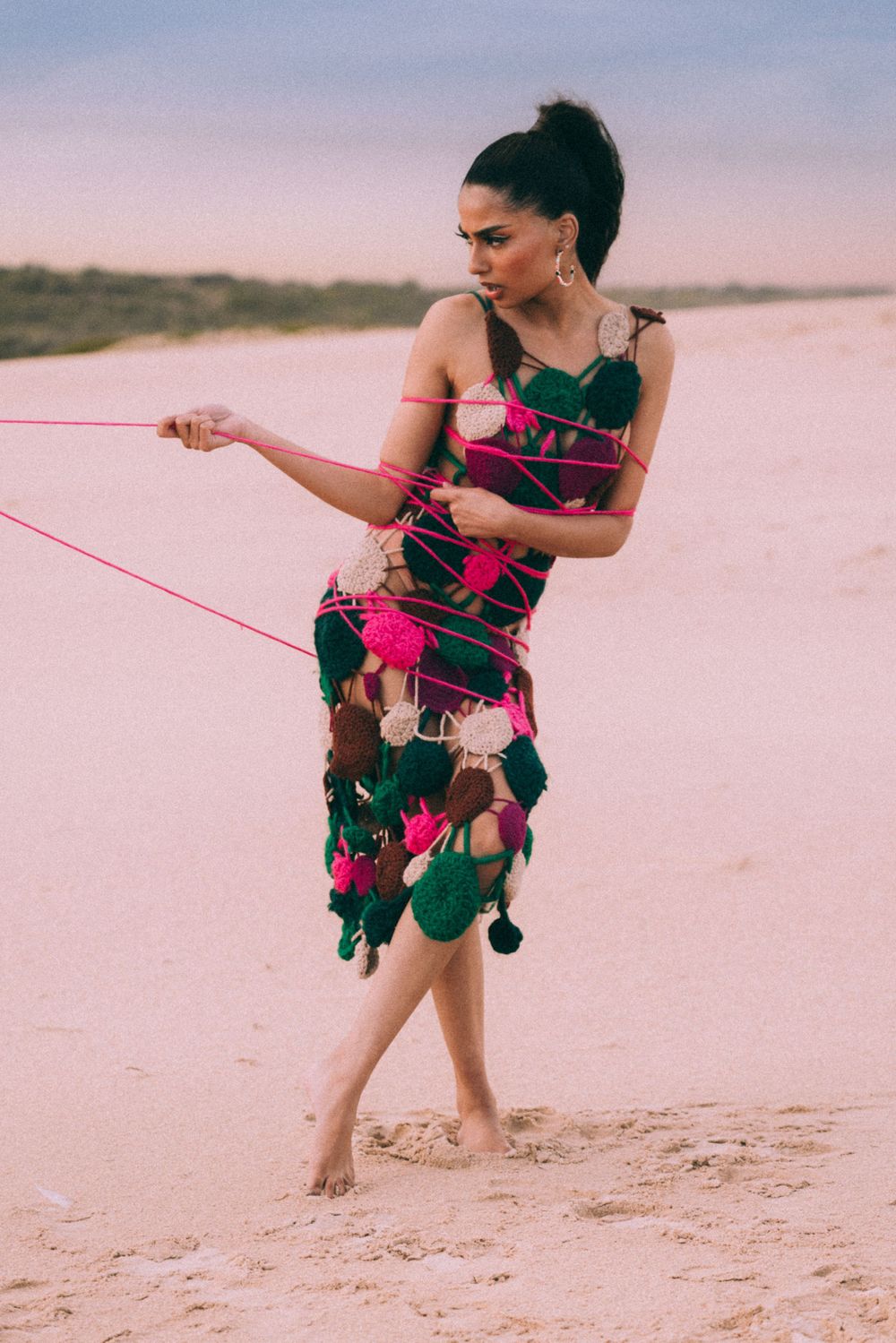 Model wearing vibrant floral dress with pink yarn details dances on sandy beach.