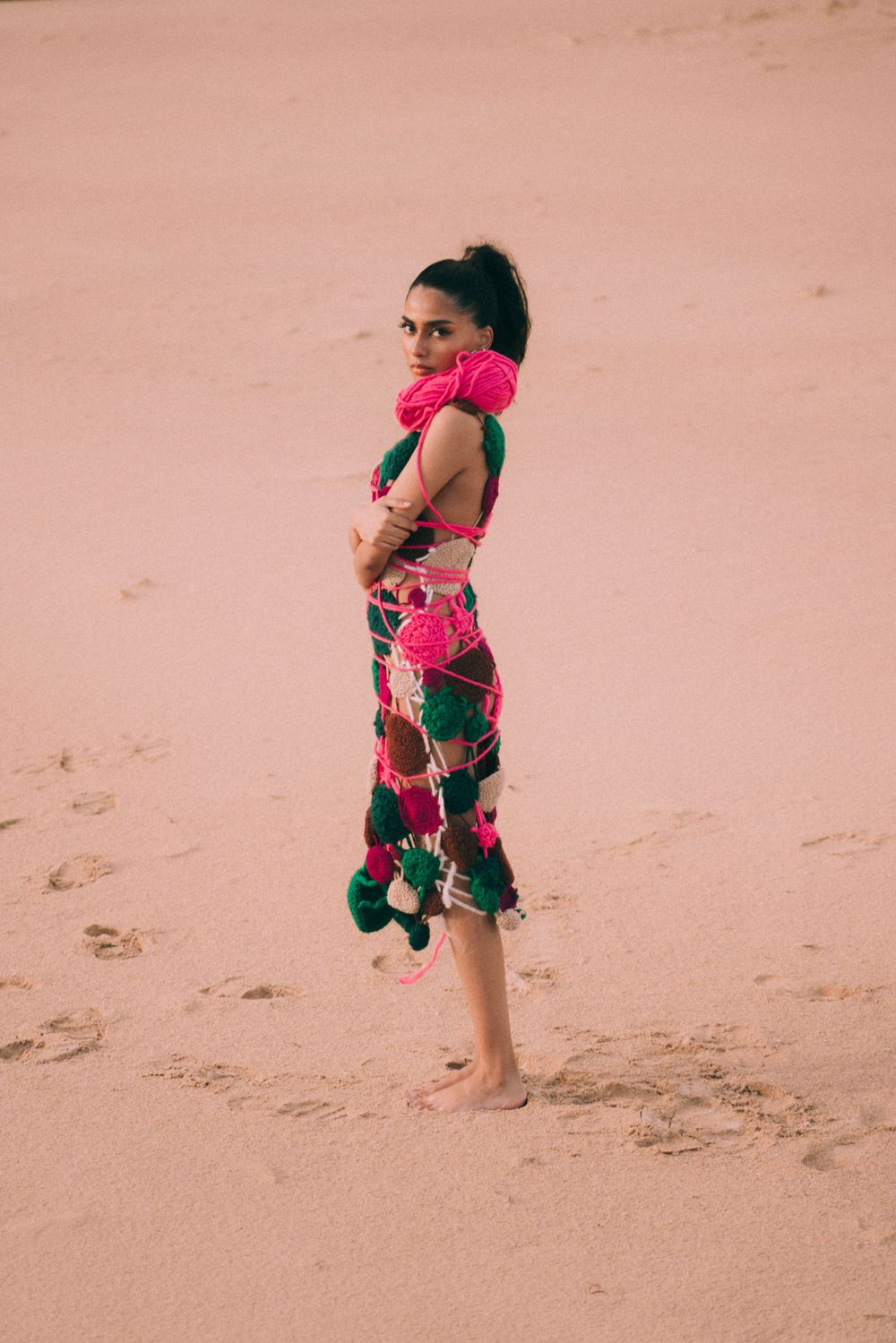 Model in colorful pom-pom adorned dress poses barefoot on pink sand dunes.