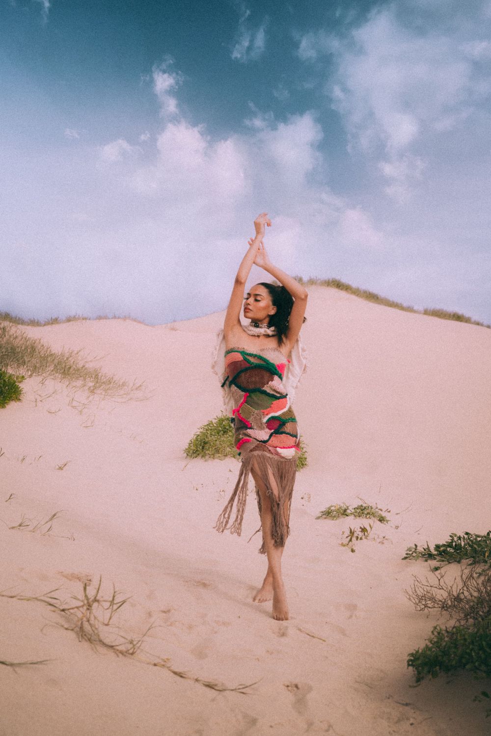 Dancing figure in patterned dress on sandy dunes against cloudy sky.