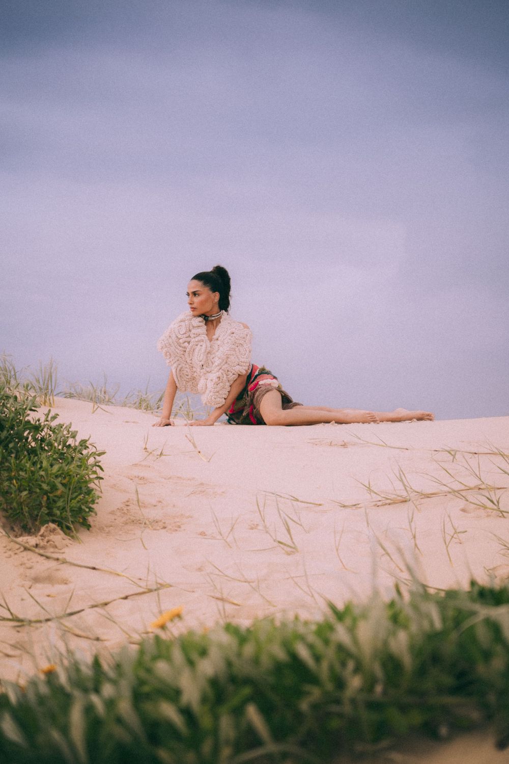 Fashion model poses gracefully on sandy dunes against dreamy purple sky at sunset.