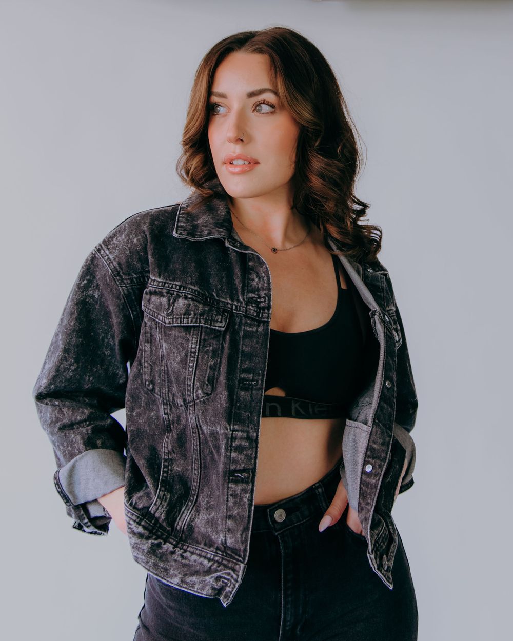 Fashion model wearing black denim jacket and crop top poses against light background.