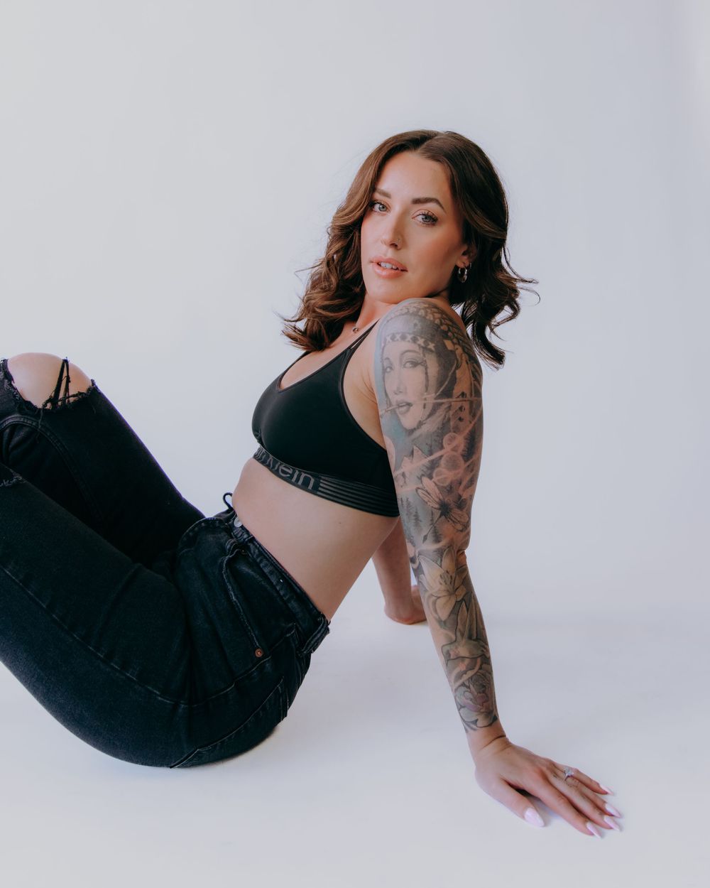 Model with tattoos poses in black outfit against minimalist white background.