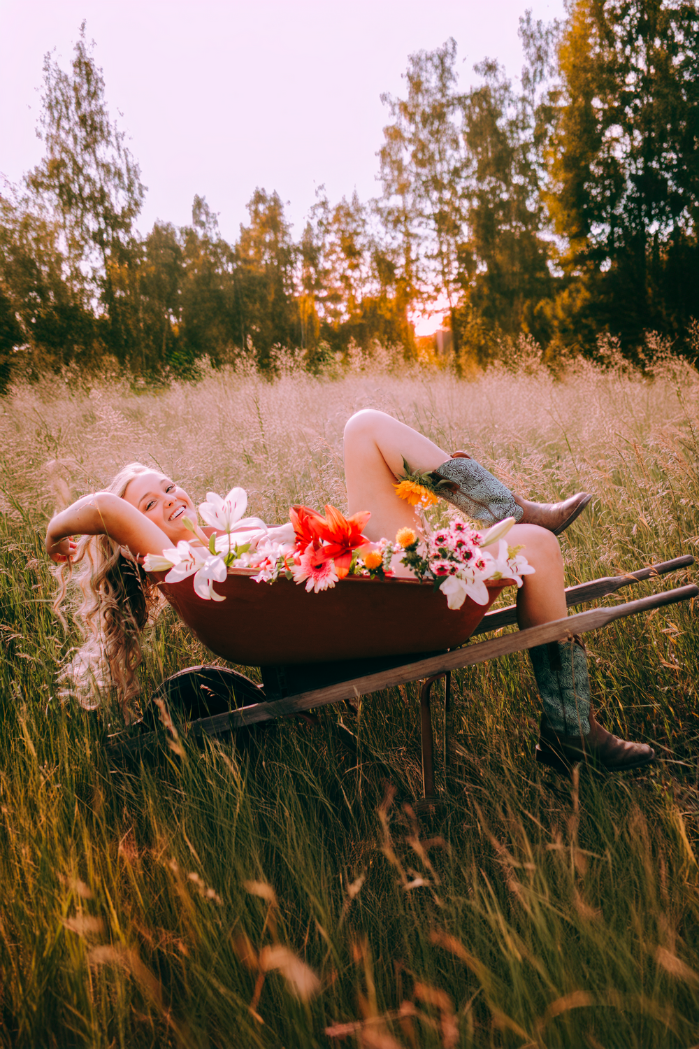 Artistic photo of person lying in wheelbarrow with flowers at sunset in field.