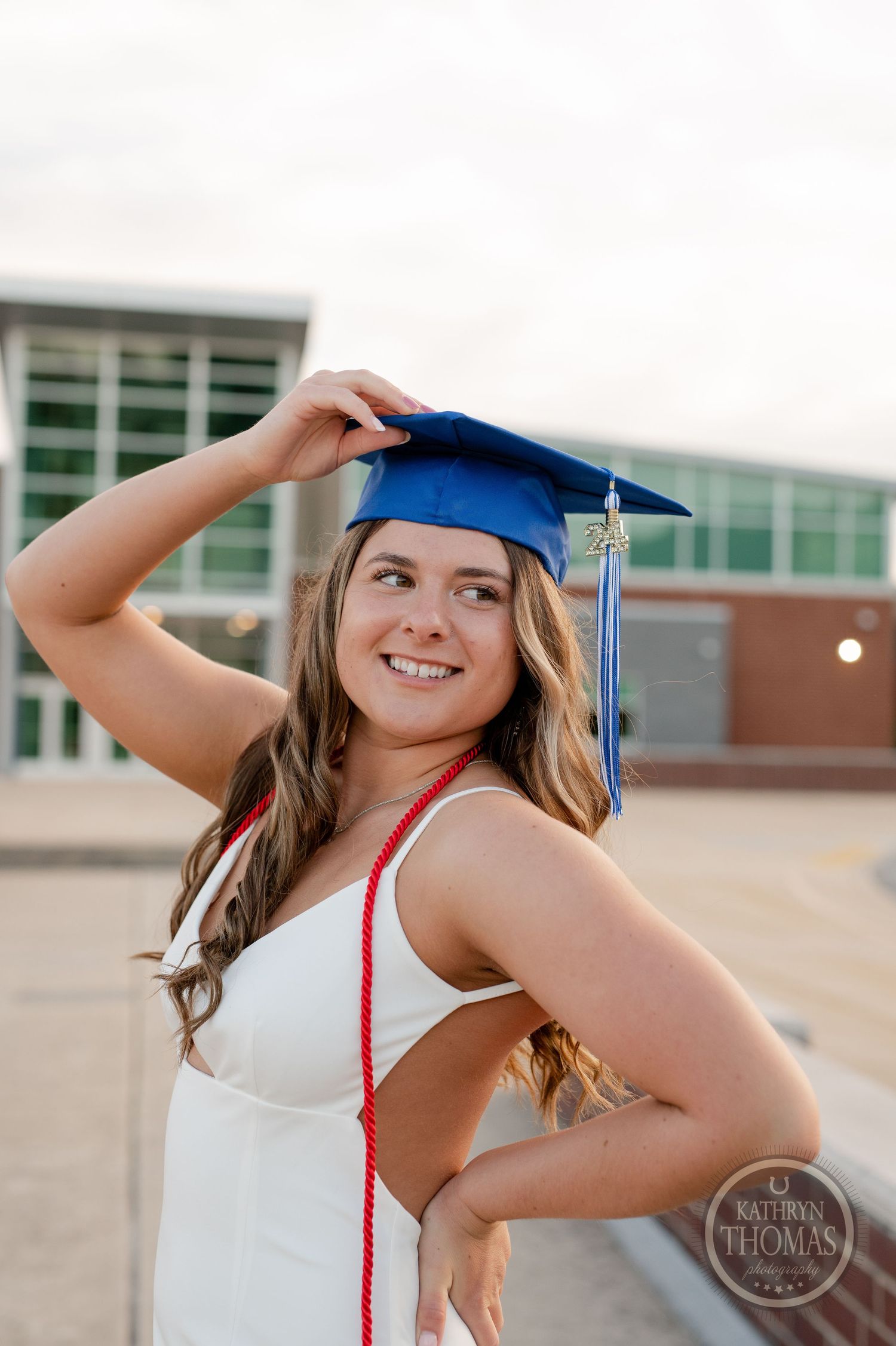 Graduate in white dress adjusts blue cap while posing for graduation photos.