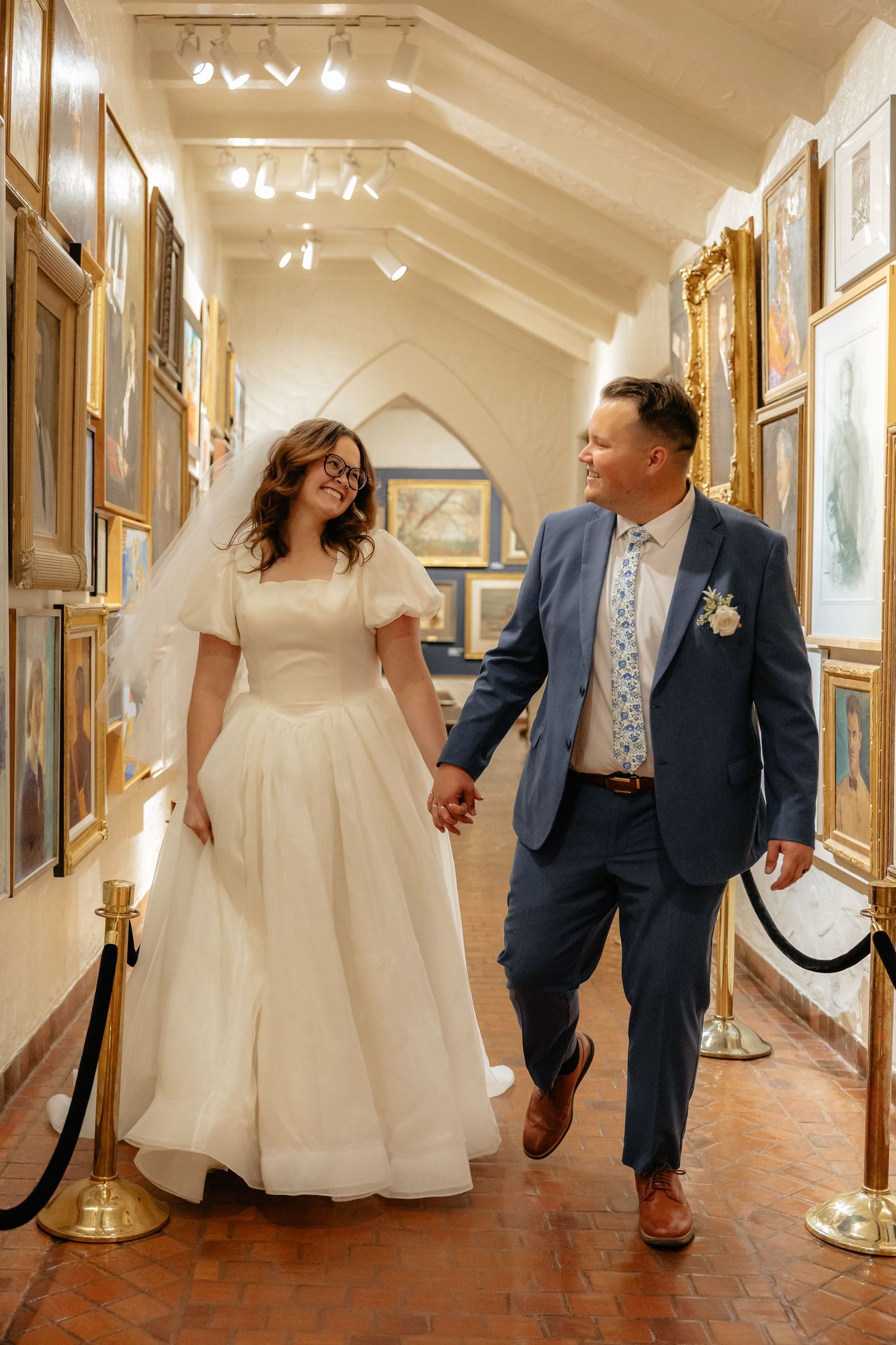 Bride in white gown and groom in blue suit walking hand in hand through an art gallery.