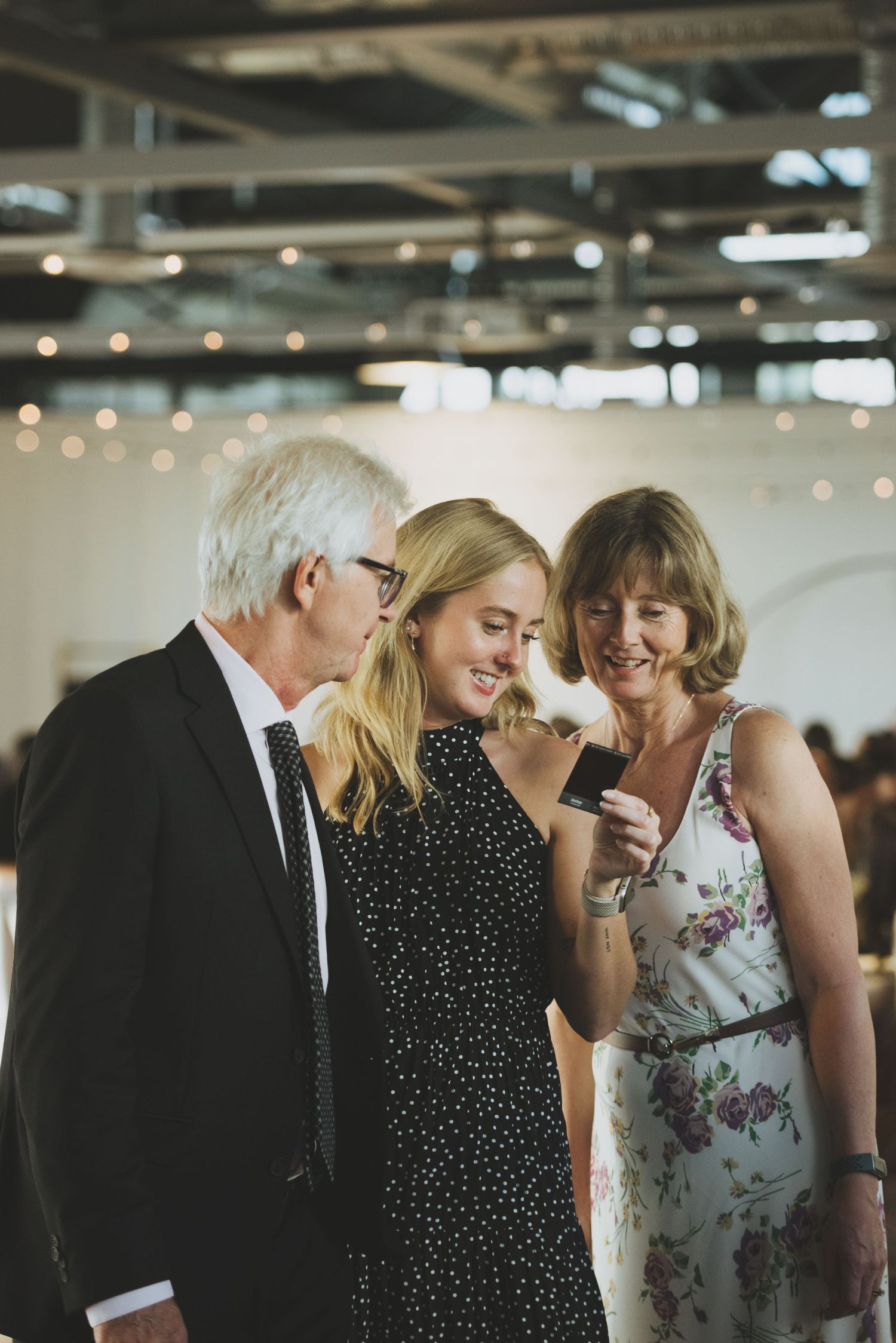 Three people share a lighthearted moment at an indoor event with twinkling lights in the background.