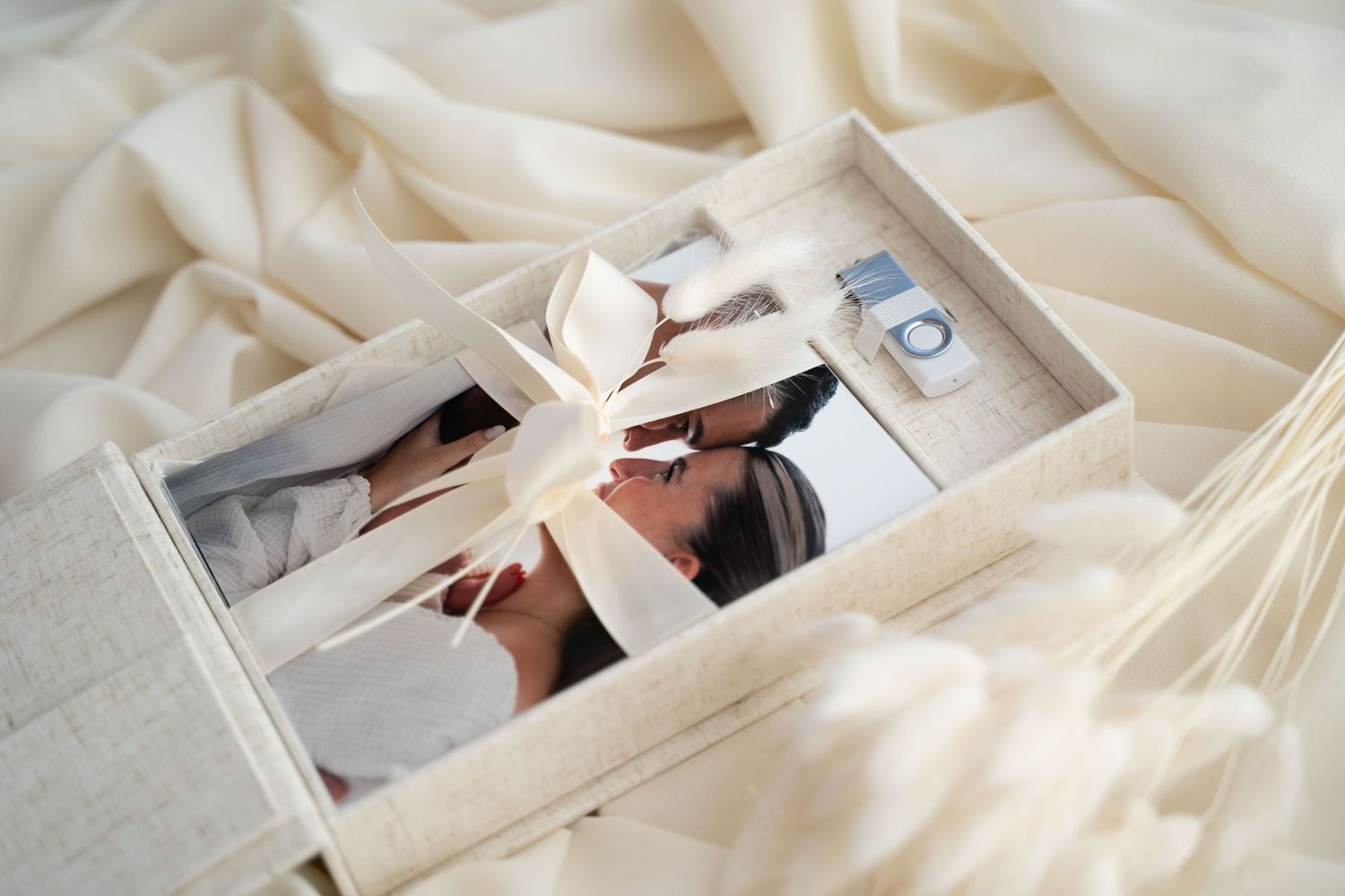 Open linen photo box with cream ribbon bow, printed photos and USB drive displayed on white silk fabric.