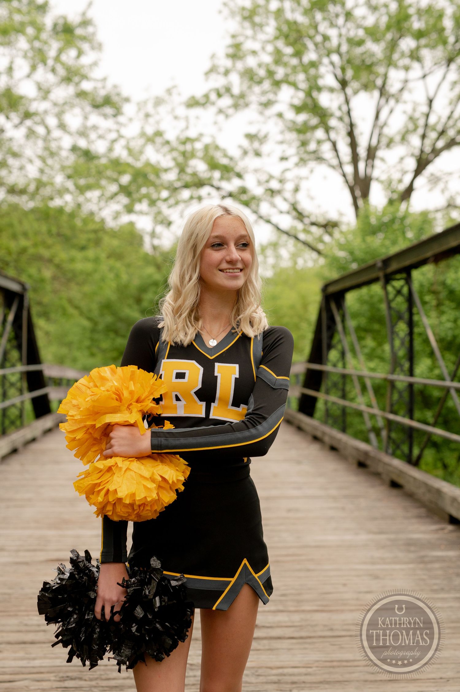 Cheerleader holds black and gold pom poms while standing on wooden bridge in park setting.