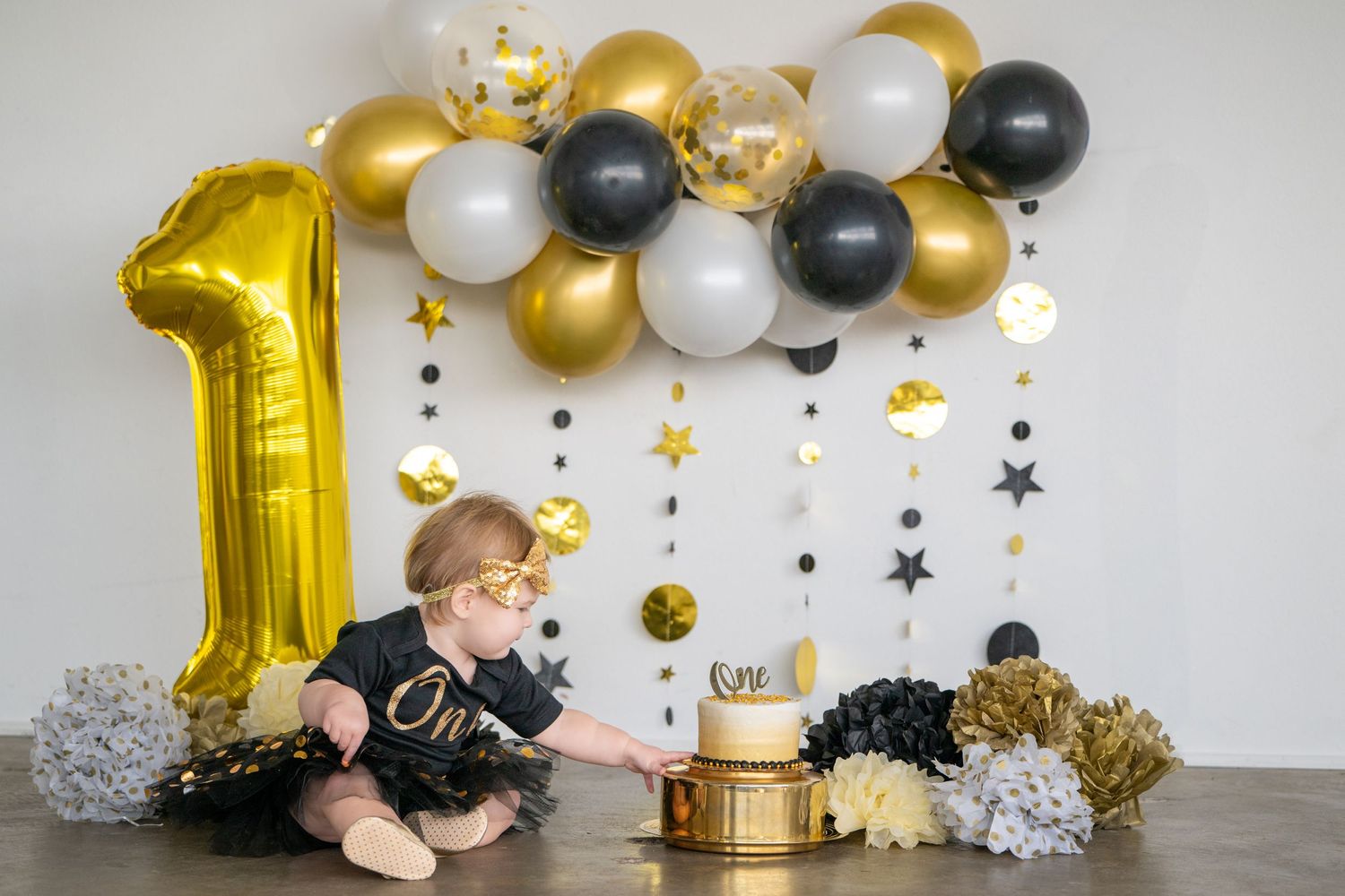A first birthday celebration setup with gold and black balloons, cake, and decorations on a wooden table.