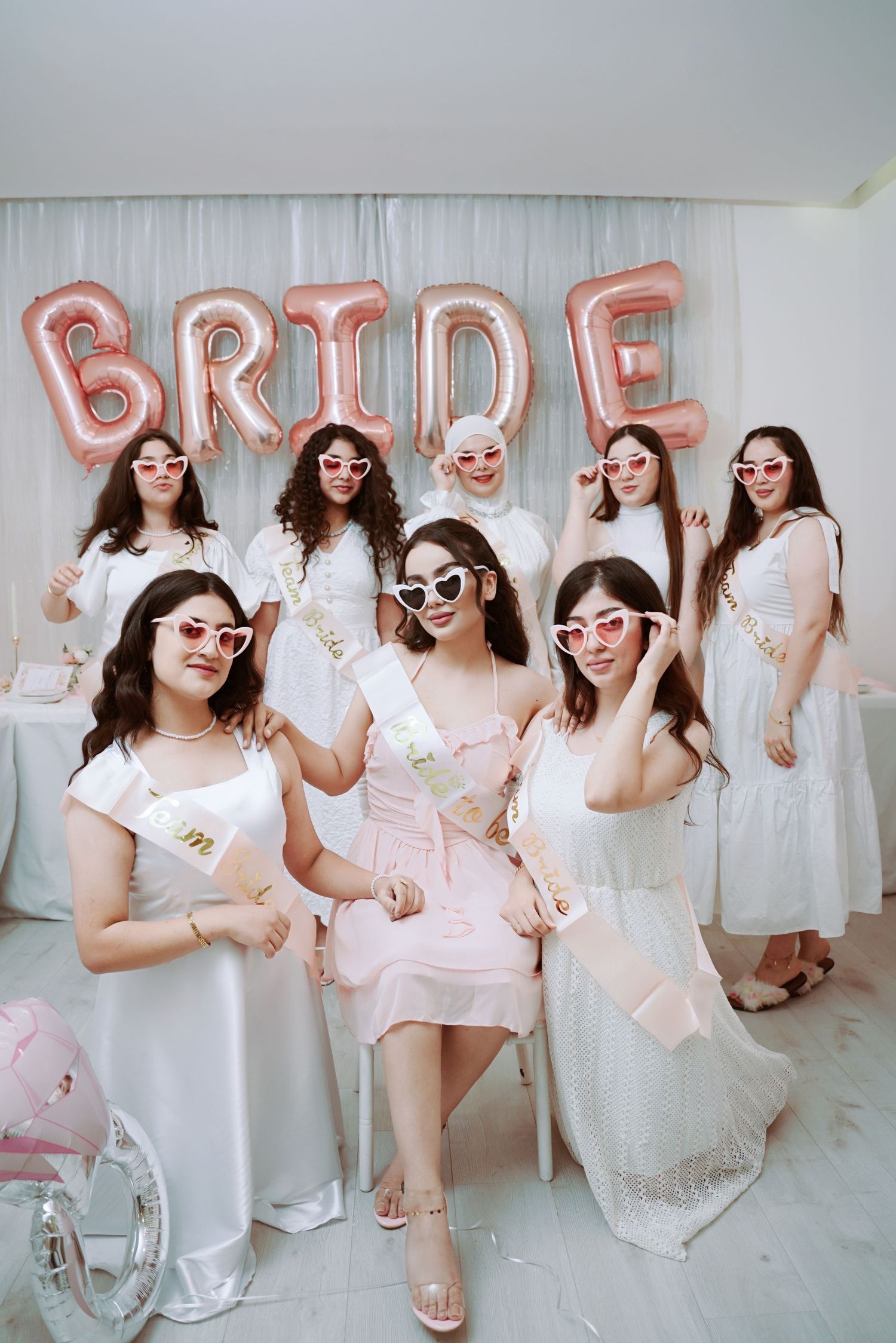 A bridal party in white dresses and sunglasses poses beneath pink balloon letters spelling BRIDE at a wedding celebration.