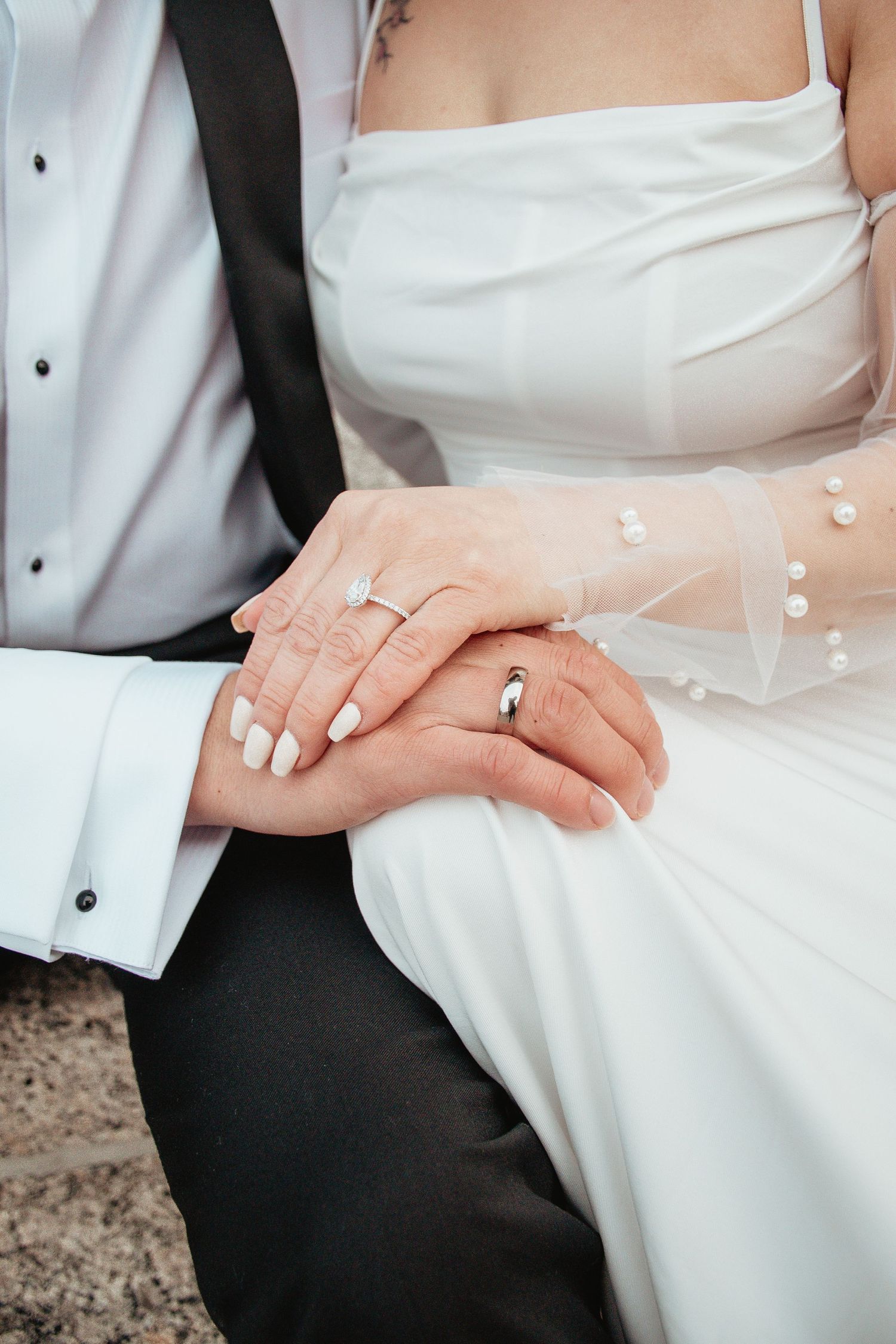 Close-up of newlyweds' hands with wedding rings against white formal attire.