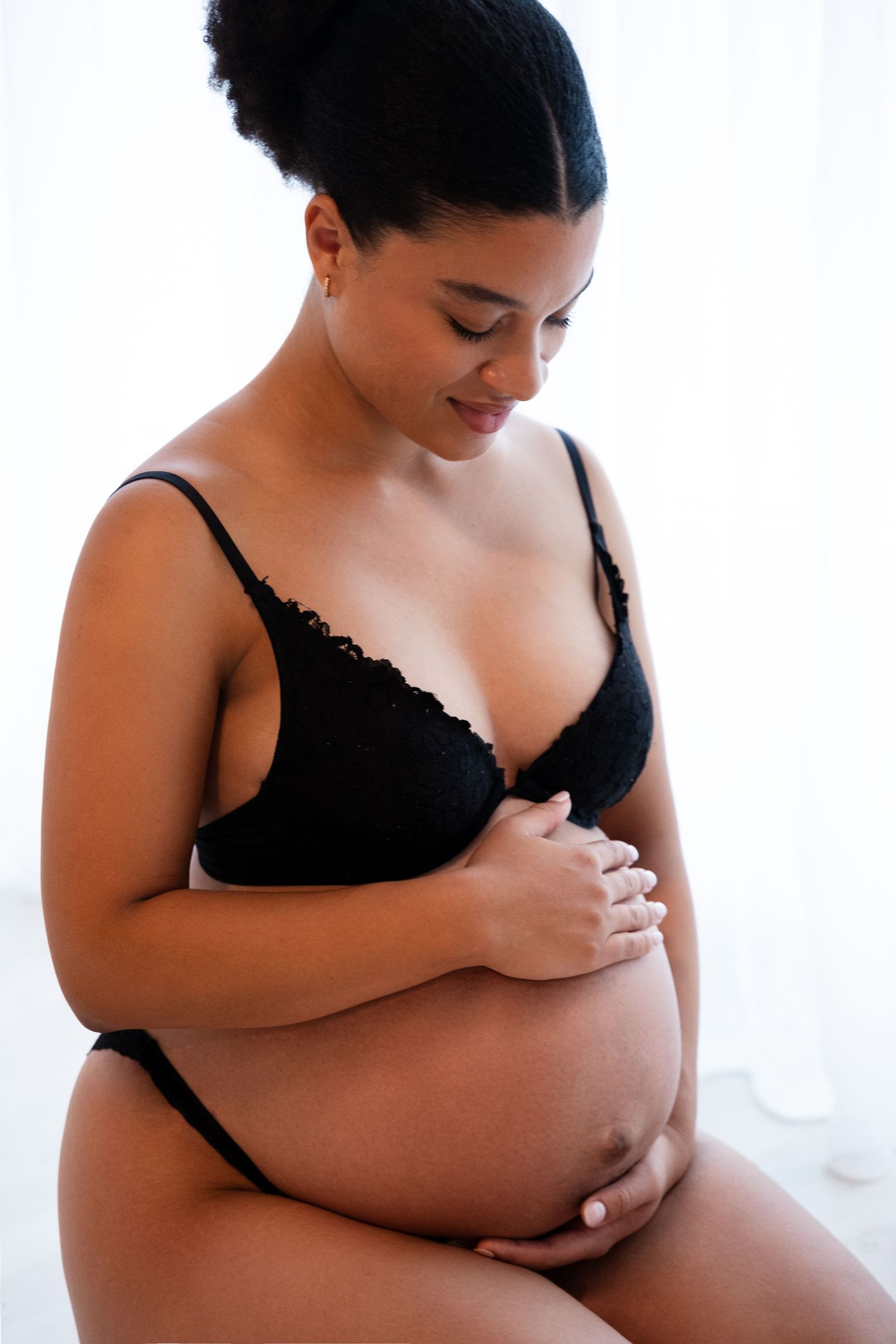 Pregnant woman in black lingerie sitting and tenderly holding her baby bump, looking down with a gentle smile.