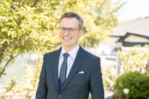 A professional in a navy suit and tie smiling outdoors on a sunny day with greenery in the background.