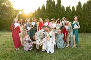 A large group in colorful dresses sits together on grass with evergreen trees in the background at sunset.