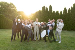 Groomsmen lined up on grassy hill toasting drinks during golden hour celebration.