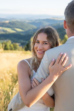A couple embraces in a golden field with rolling hills and mountains in the background during sunset.