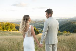 A couple stands together in a golden meadow at sunset with rolling hills in the background.