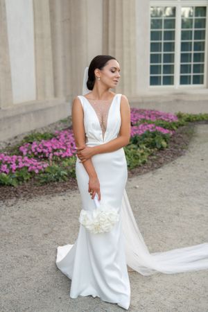Elegant white wedding dress with plunging neckline and flowing train against pink floral garden backdrop.