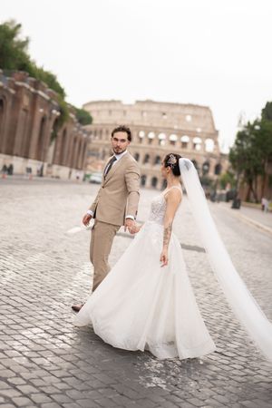 A couple in wedding attire walking together on cobblestone streets near the Colosseum in Rome, Italy.